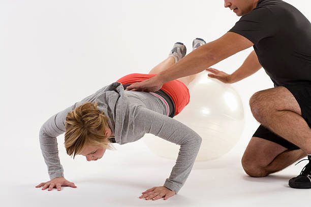 A female patient performs a stability ball push-up exercise with assistance from a chiropractor or physical therapist, focusing on core stabilization and proper form.