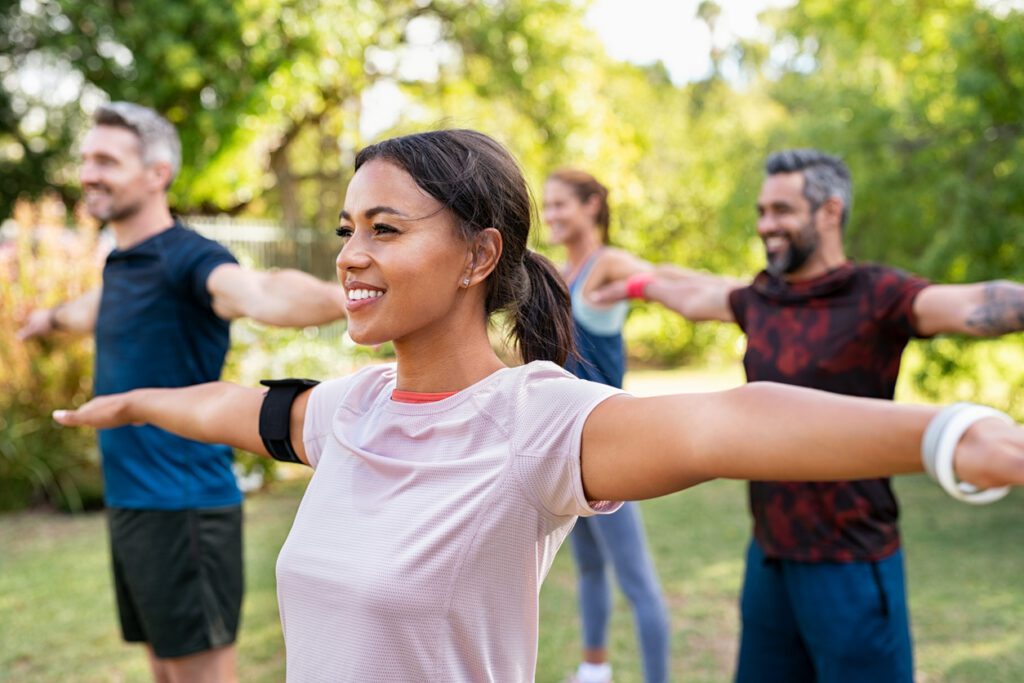 A diverse group of four adults smiling and stretching their arms out to the sides during an outdoor fitness class in a park.